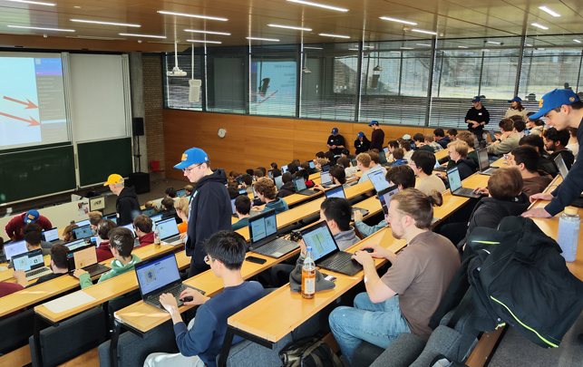 Gruppe Studierender in Hörsaal mit Laptops, Vorlesung und Referenten an der Wand.